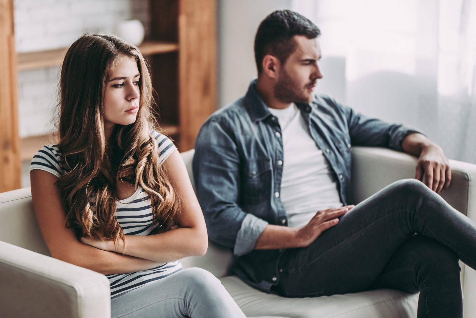  Couple sitting on a couch upset and facing away from each other. She is crossing her arms on the left and he is crossing his legs on the right